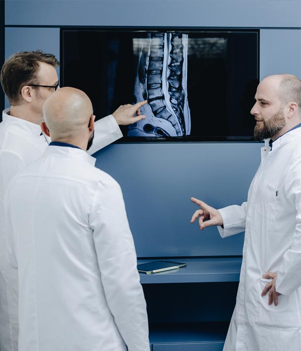 Fachärzte für Neurochirurgie stehen vor einem MRT-Bild einer Wirbelsäule. Einer von ihnen zeigt auf das Bild.